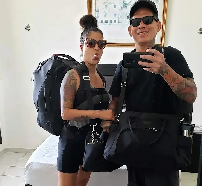 MJ and me in a mirror selfie, both dressed in black, carrying a backpack, a duffel and two shoulder bags between us, in a small room with a made bed behind us.